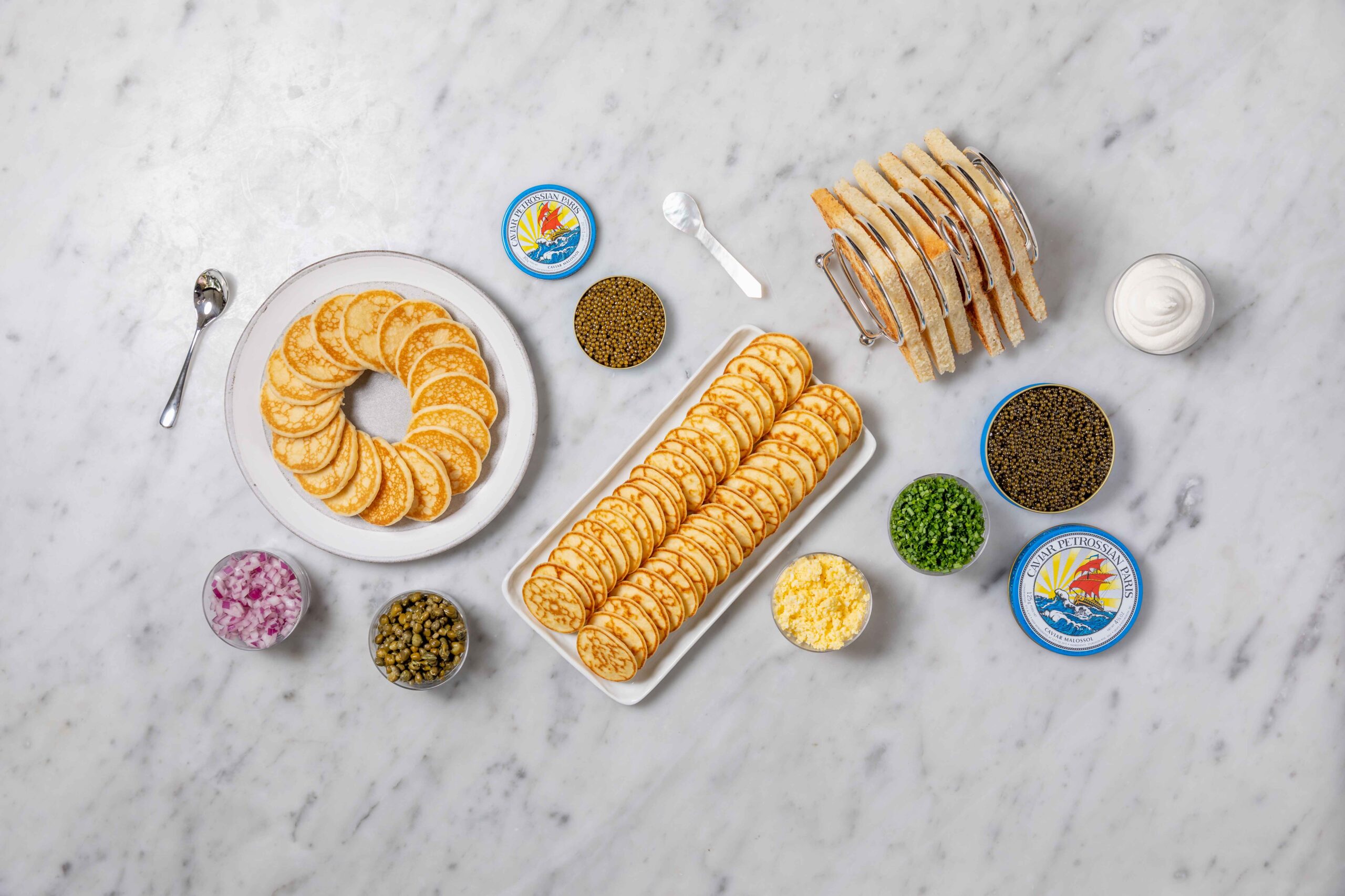 Plates of blinis and toasts, surrounded by tins of caviar, creme fraiche and pearl spoons on a white counter top