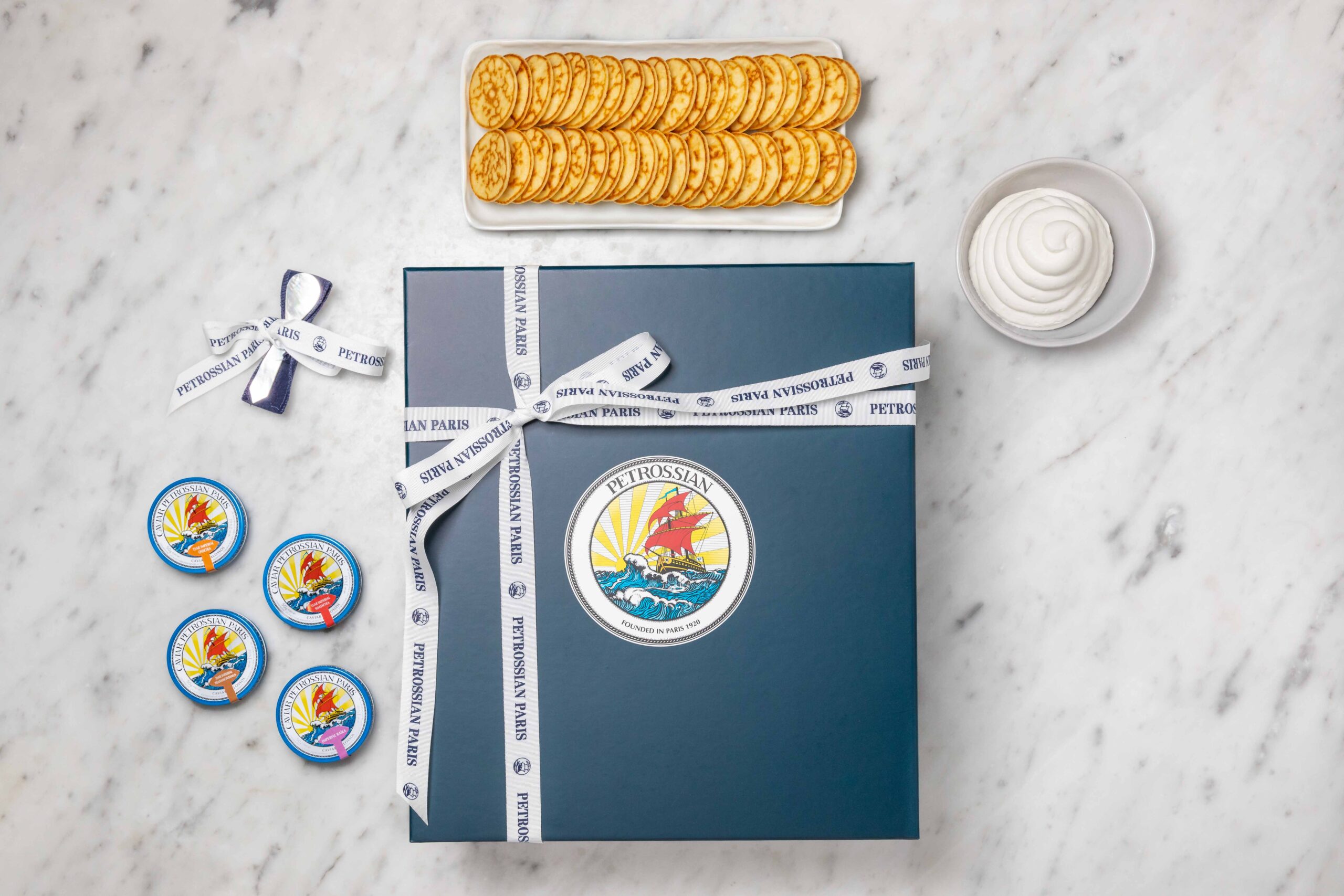 a navy blue Petrossian gift box with white ribbons and many blue tins of caviar and blinis, all sitting on white marble counter top