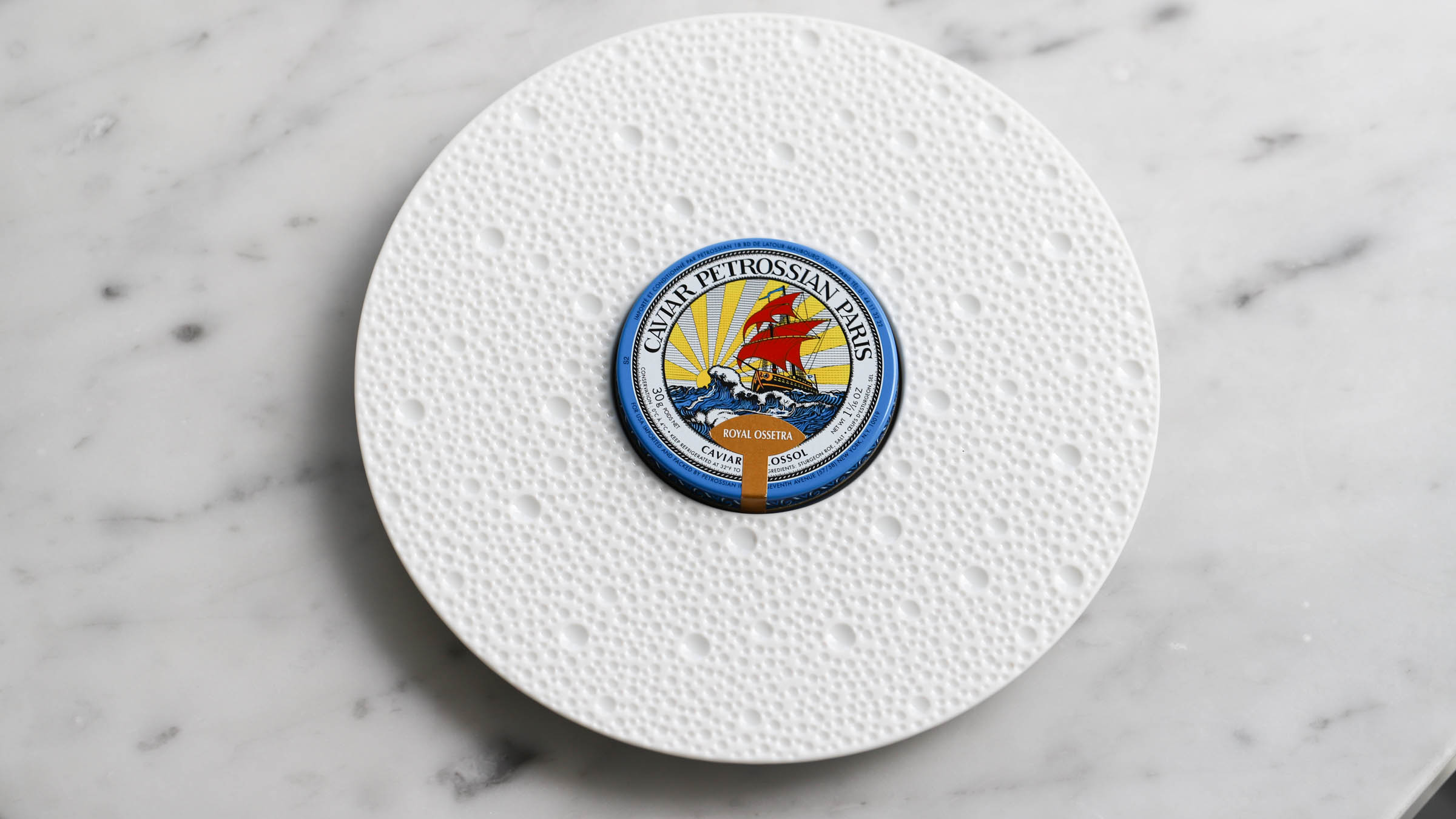 a white porcelain plate on a white marble counter top. on the plate sits a blue unopened tin of caviar.