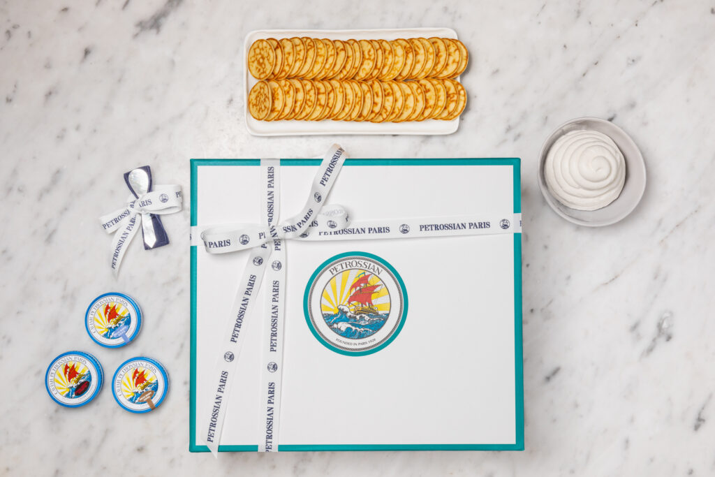 a white Petrossian gift box with teal accents on a white marble surface, surrounded by blinis and tins of caviar
