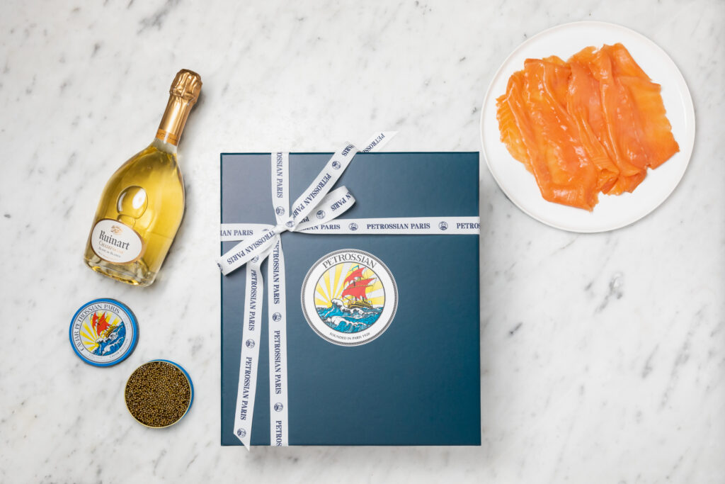a navy blue Petrossian gift box on a white countertop surrounded smoked salmon, caviar and a bottle of champagne