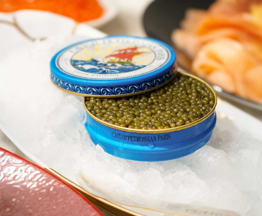 a blue int of Petrossian caviar sitting in crushed ice. the caviar is plump and green and glistens in the light.