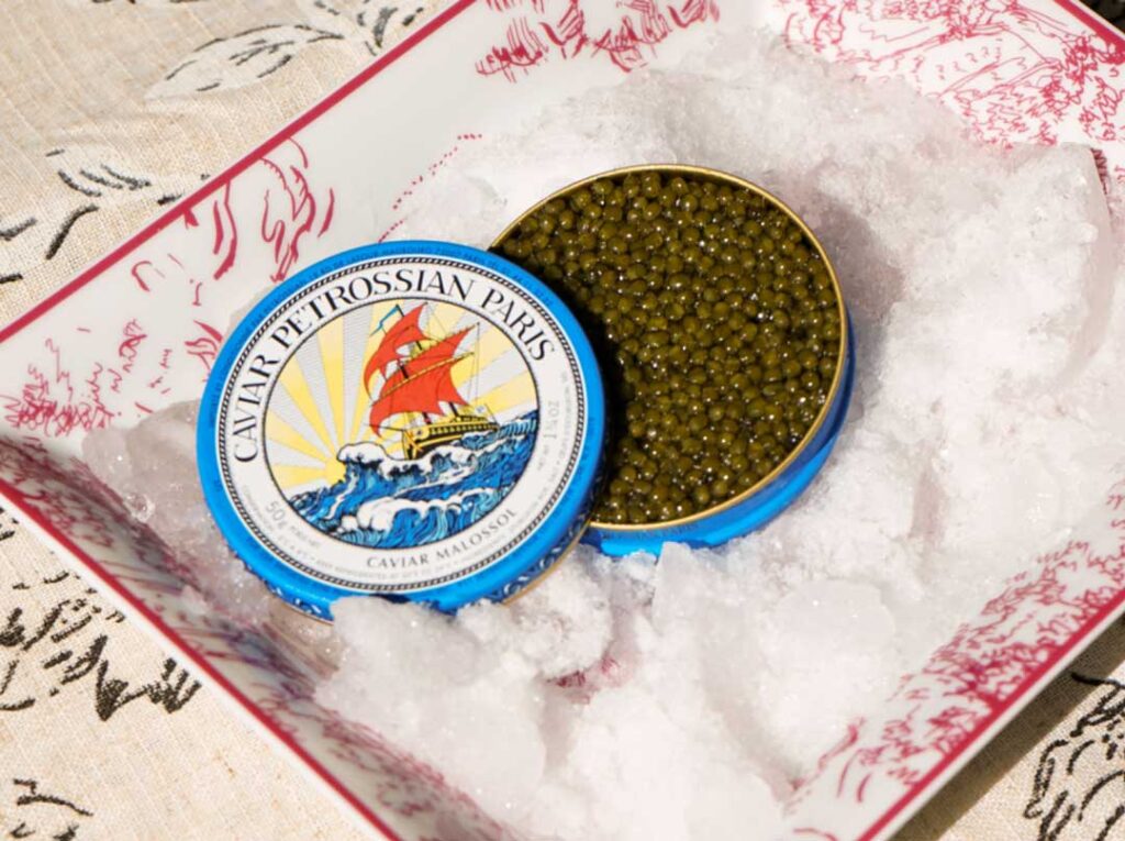 a blue tin of Petrossian caviar with the Petrossian logo on the lid. the tin sits in crushed ice and the brown-green caviar reflects the dramatic light.